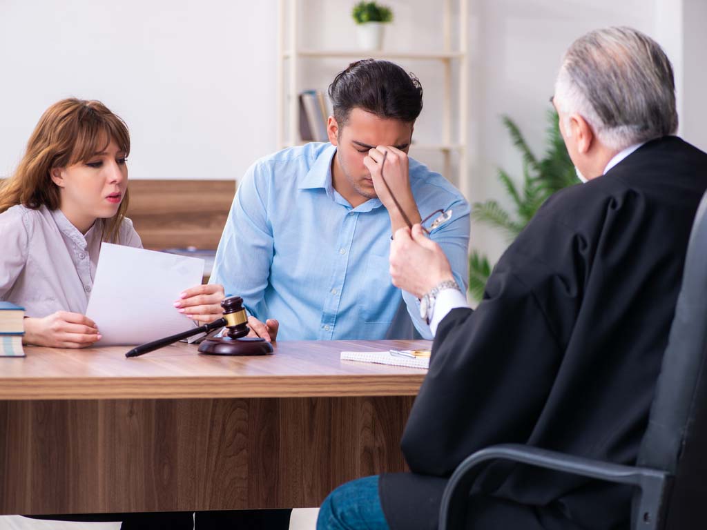 woman sitting next to man holding the bridge of his nose as judge talks to him